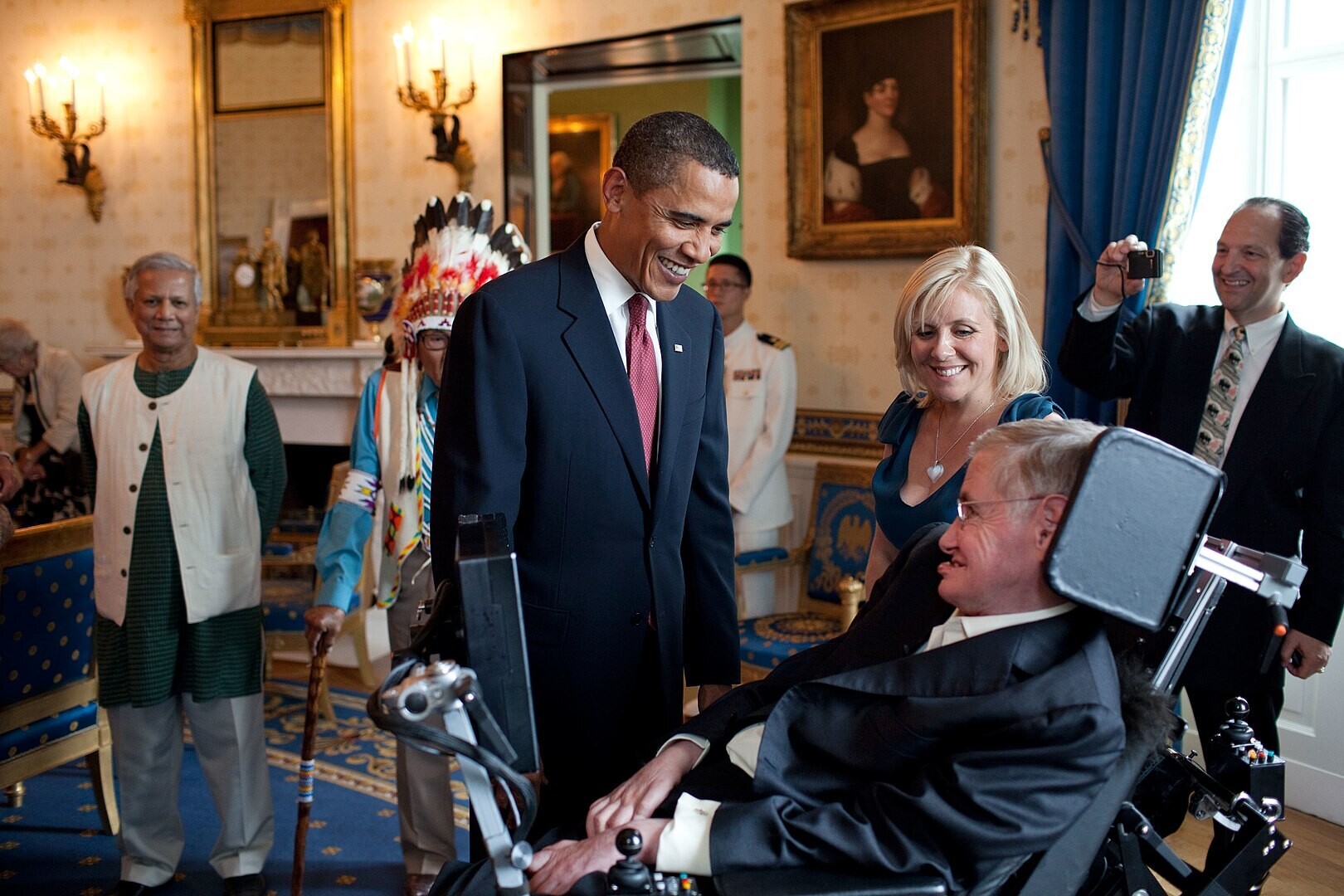Barack Obama standing in front of Stephen Hawking’s wheelchair talking to him. They are surrounded by other award winners.