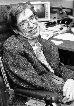 Hawking, dressed in shirt and suit coat, sitting in a wheelchair. In the background a desk and computer are visible.