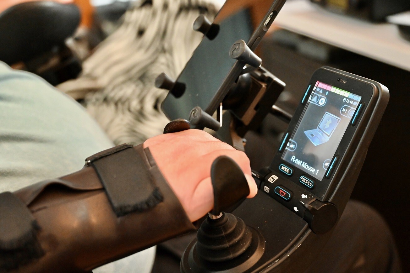 La main droite de Cyrill manie le joystick. Un petit écran indique que le fauteuil roulant et l’ordinateur sont connectés.
