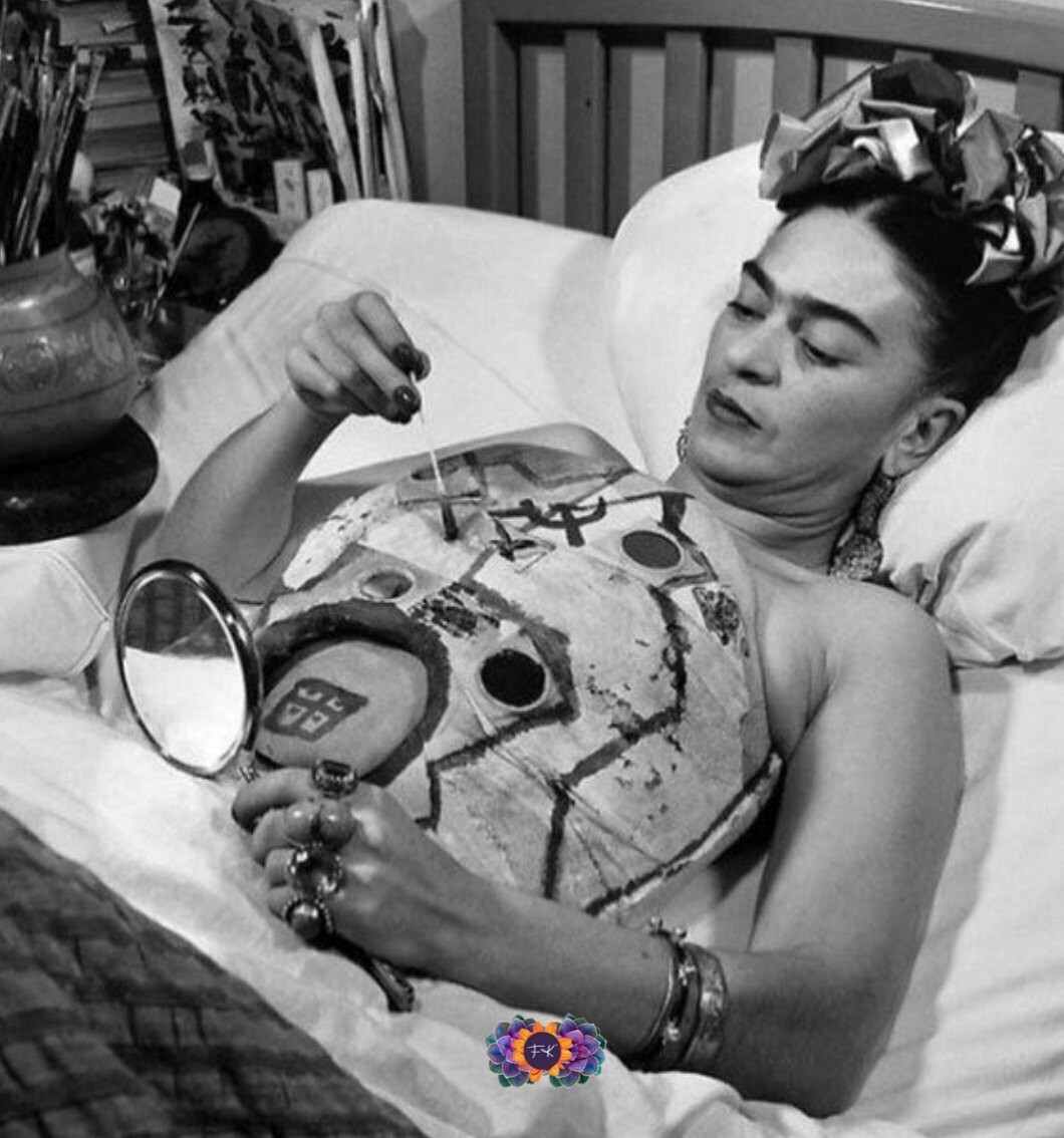Frida Kahlo liegt auf dem Rücken im Bett. Sie hält einen Spiegel in der Hand und bemalt ihr Korsett.