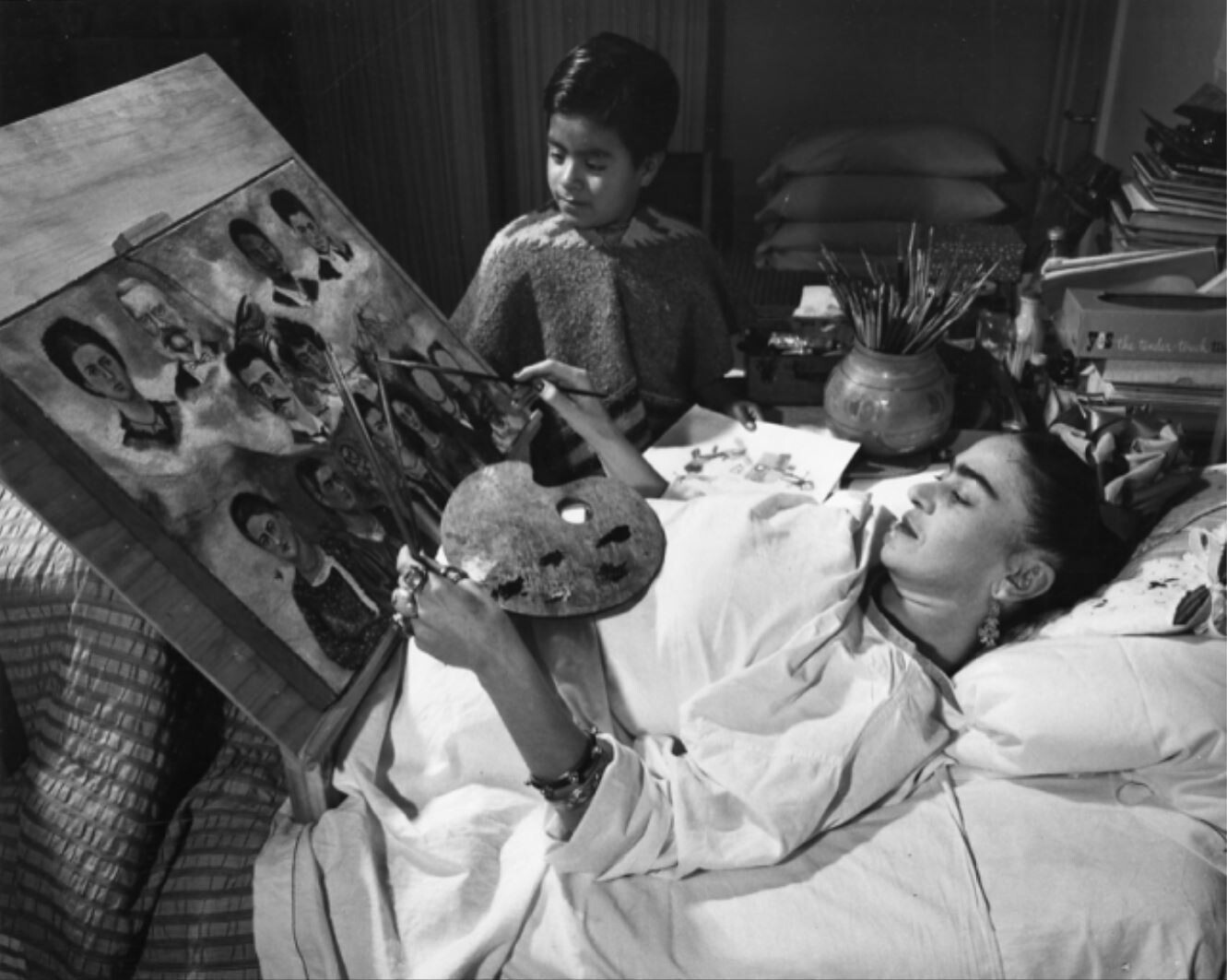 Frida Kahlo lying on her back in bed. In front of her a wooden structure with a canvas is installed, a color palette on her belly.