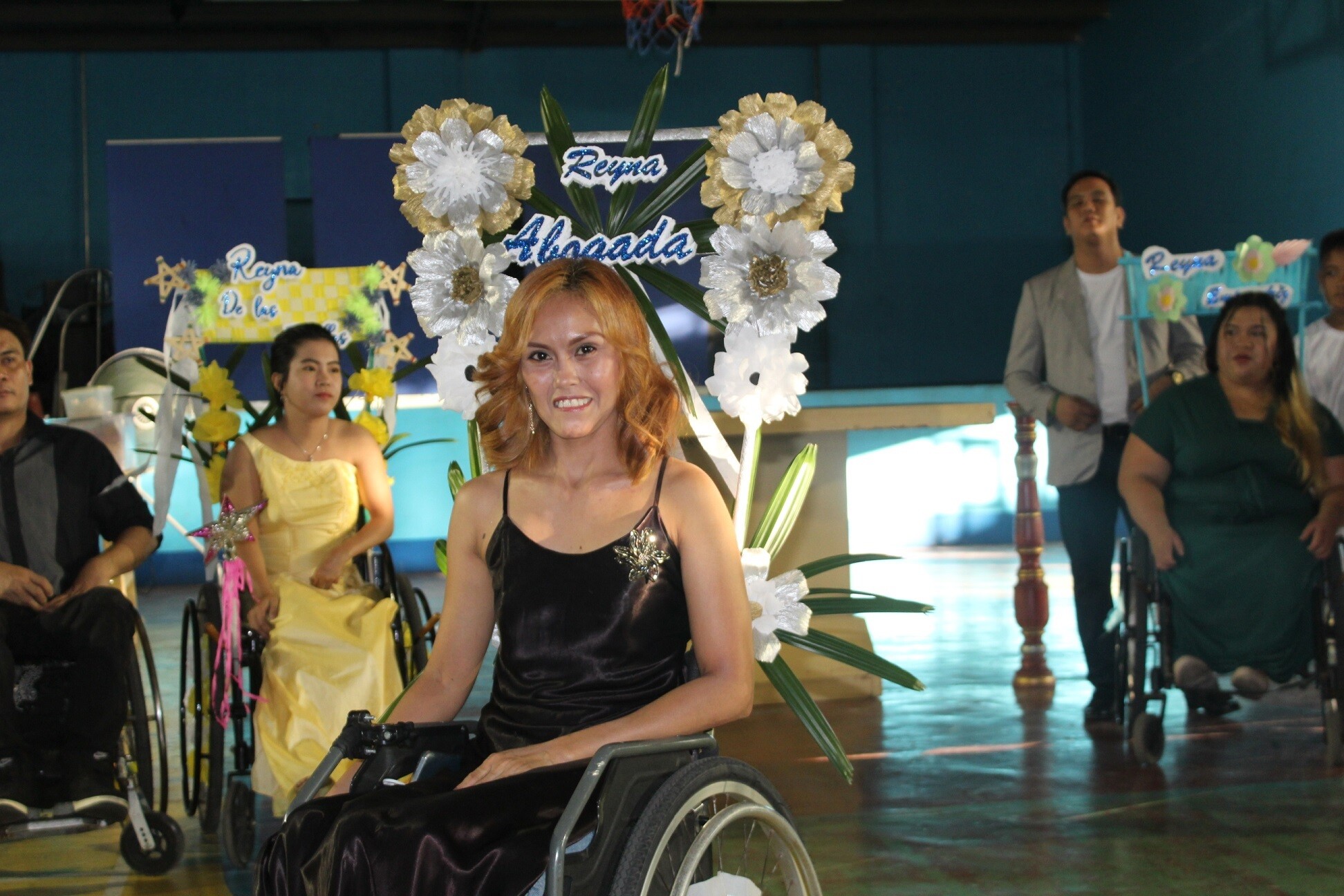 Jobelle, with a black dress and with red-coloured hair, at a party. Her wheelchair is decorated with flowers. Other wheelchair users are in the background.