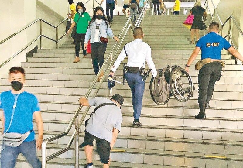 Two guards are walking up on stairs while holding a wheelchair. Several steps behind them, the wheelchair owner is struggling to walk up the stairs.