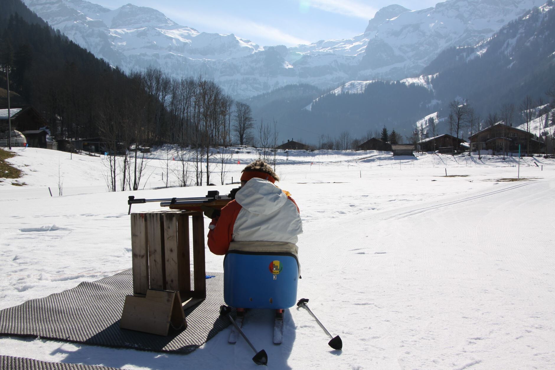 A man in a cross-country sled rests his rifle on a wooden box to aim at the biathlon targets.