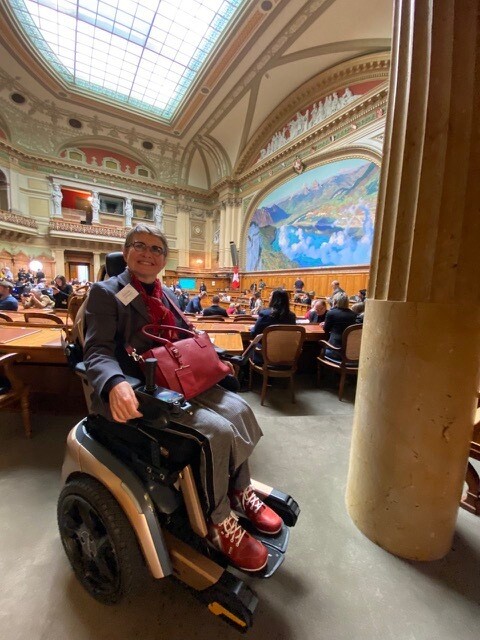 Anne Hägler poses in her wheelchair in the National Council chamber, the rows of lecterns stretch out behind her.