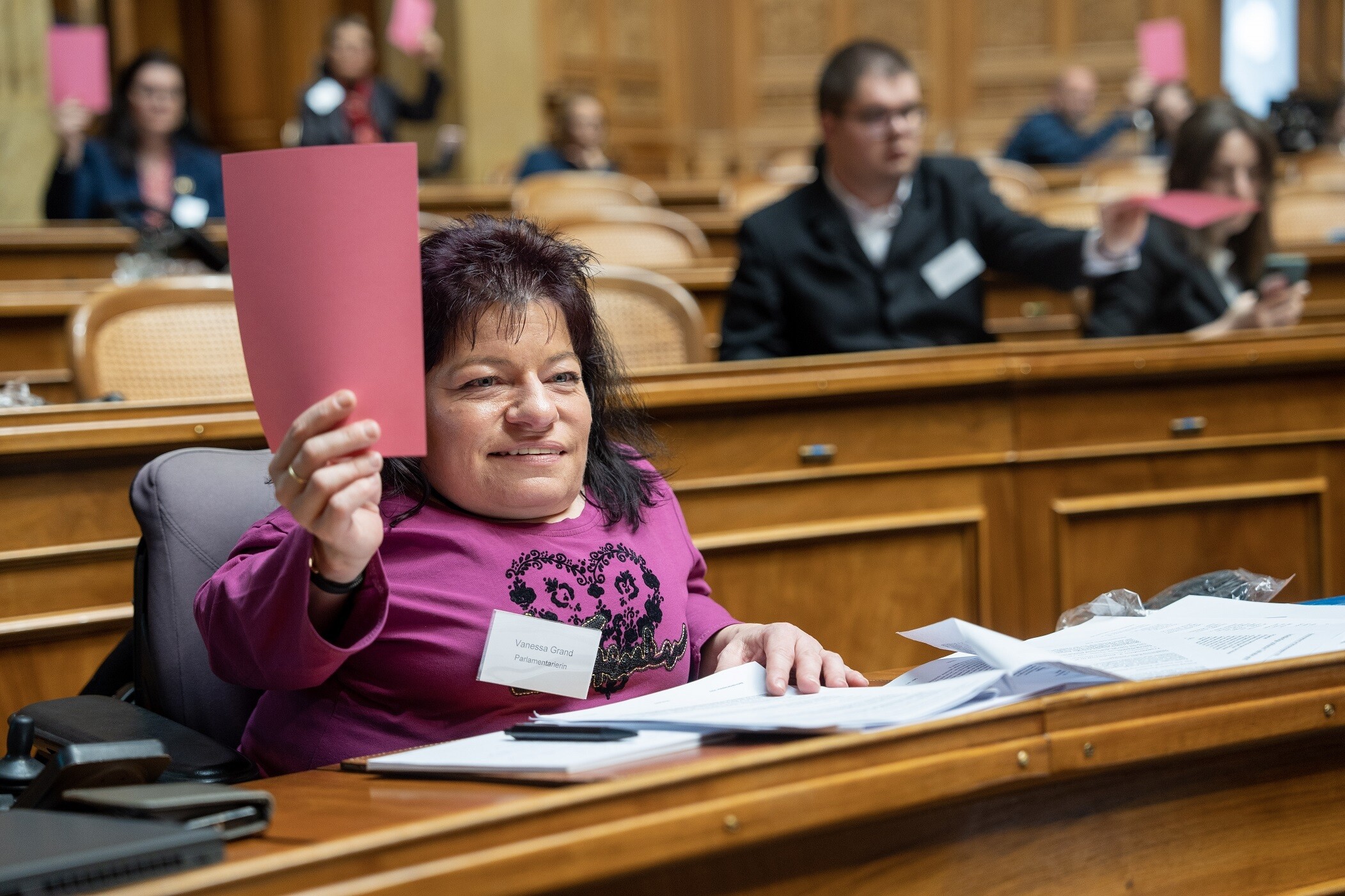 Vanessa Grand siede dietro una scrivania nella sala del Consiglio Nazionale e solleva in aria la scheda elettorale rossa con la mano destra.
