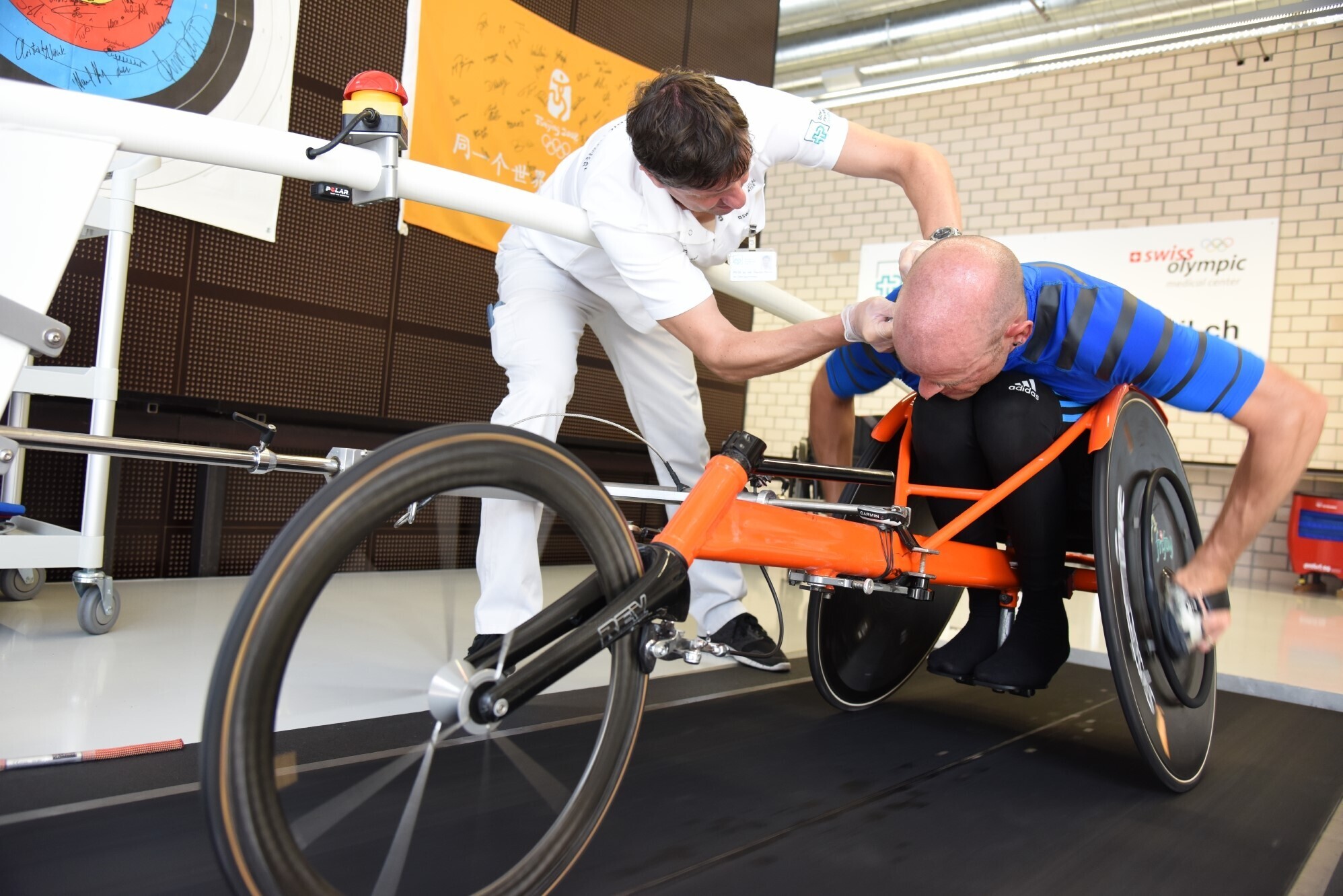 A man in a race wheelchair on a treadmill. Next to him, Claudio Perret is bending over to attach a measuring tool to the man's ear. They are in a sports laboratory room.