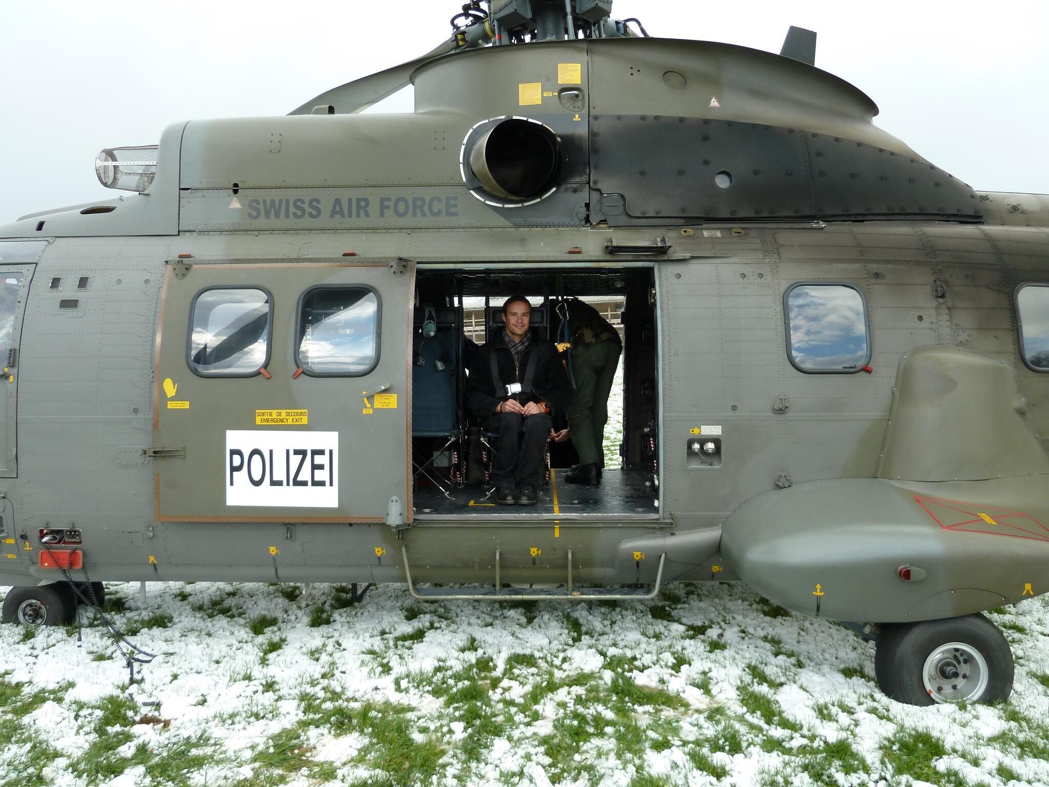 Der Polizeihelikopter steht auf einer schneebedeckten Wiese, die Schiebetüre ist offen. Im Helikopter sitzt Marcel Hug, mit Gurten gesichert.