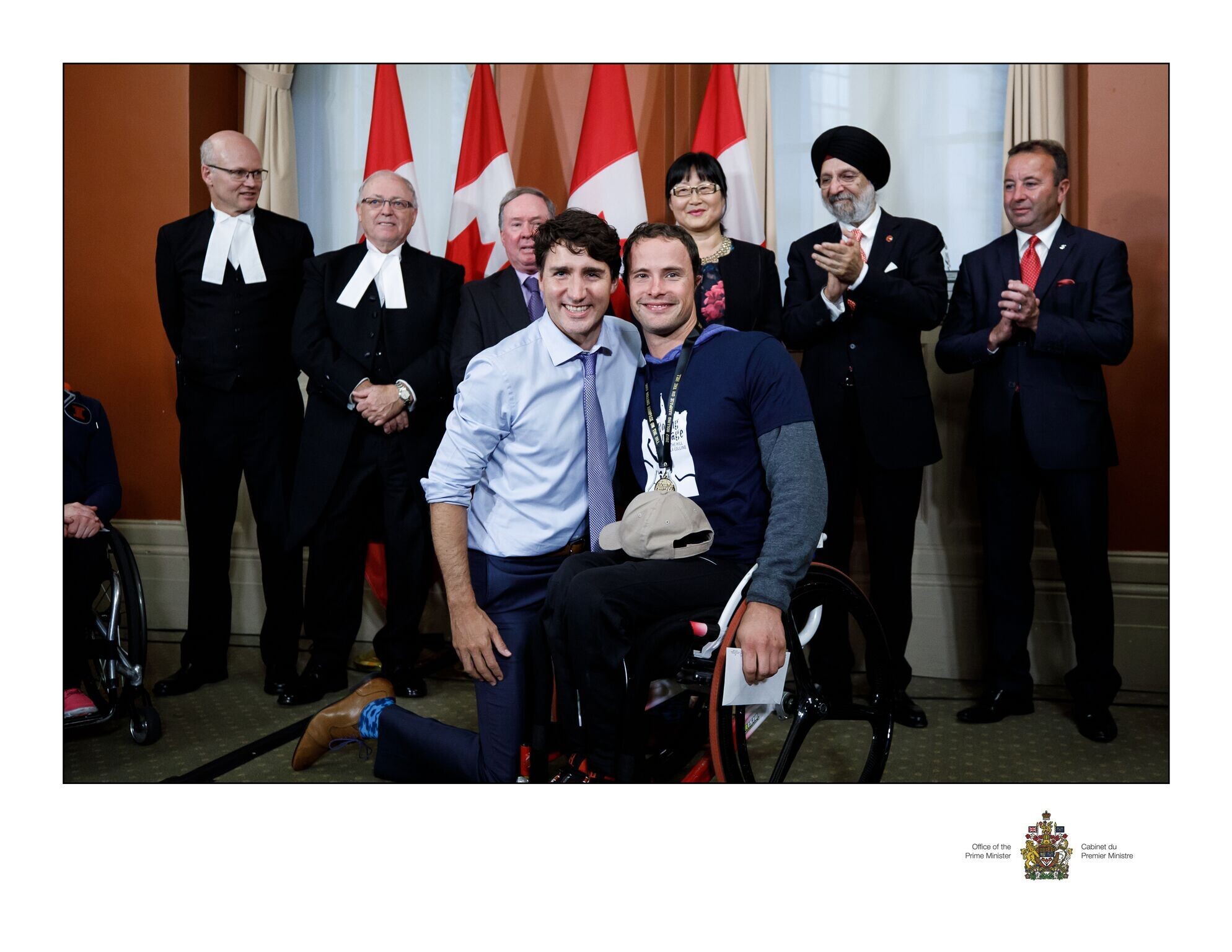Der kanadische Premierminister Justin Trudeau kniet links neben dem Rollstuhlsportler Marcel Hug für das Foto. Im Hintergrund stehen sechs applaudierende Staatsangestellte.