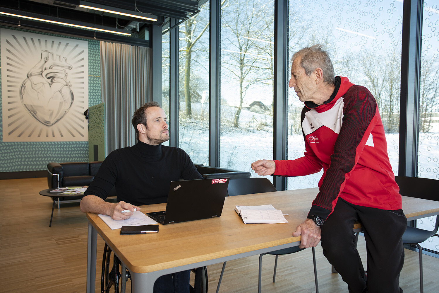 Links sitzt Marcel Hug in einem schwarzen Rollkragenpullover an einem Tisch, vor ihm Laptop und Block. Rechts sitzt sein Trainer Paul Odermatt in roter Trainerjacke lässig auf dem Tisch. Die beiden unterhalten sich.
