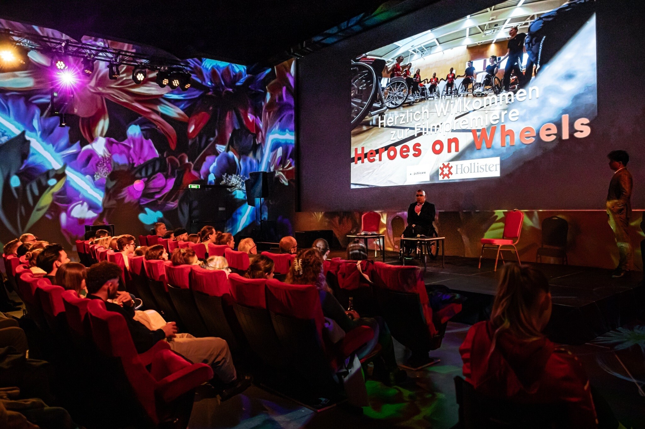 Zuschauer sitzen auf roten Kinosesseln, die Wand im Hintergrund ist farbig beleuchtet, auf der Bühne hält Dominik Rüedi ein Mikrofon in der Hand. Über ihm ein grosses Bild, auf dem die Frauen-Nationalmannschaft zu sehen ist, darunter steht «Herzlich Willkommen zur Filmpremiere Heroes on Wheels».