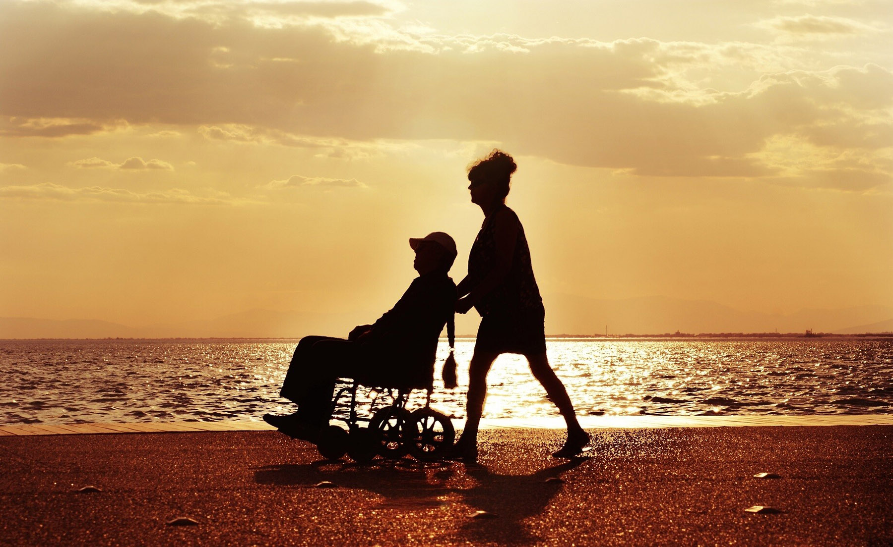 Eine Pflegefachfrau begleitet einen Mann im Rollstuhl am Strand, im Hintergrund das Meer und der Sonnenuntergang.