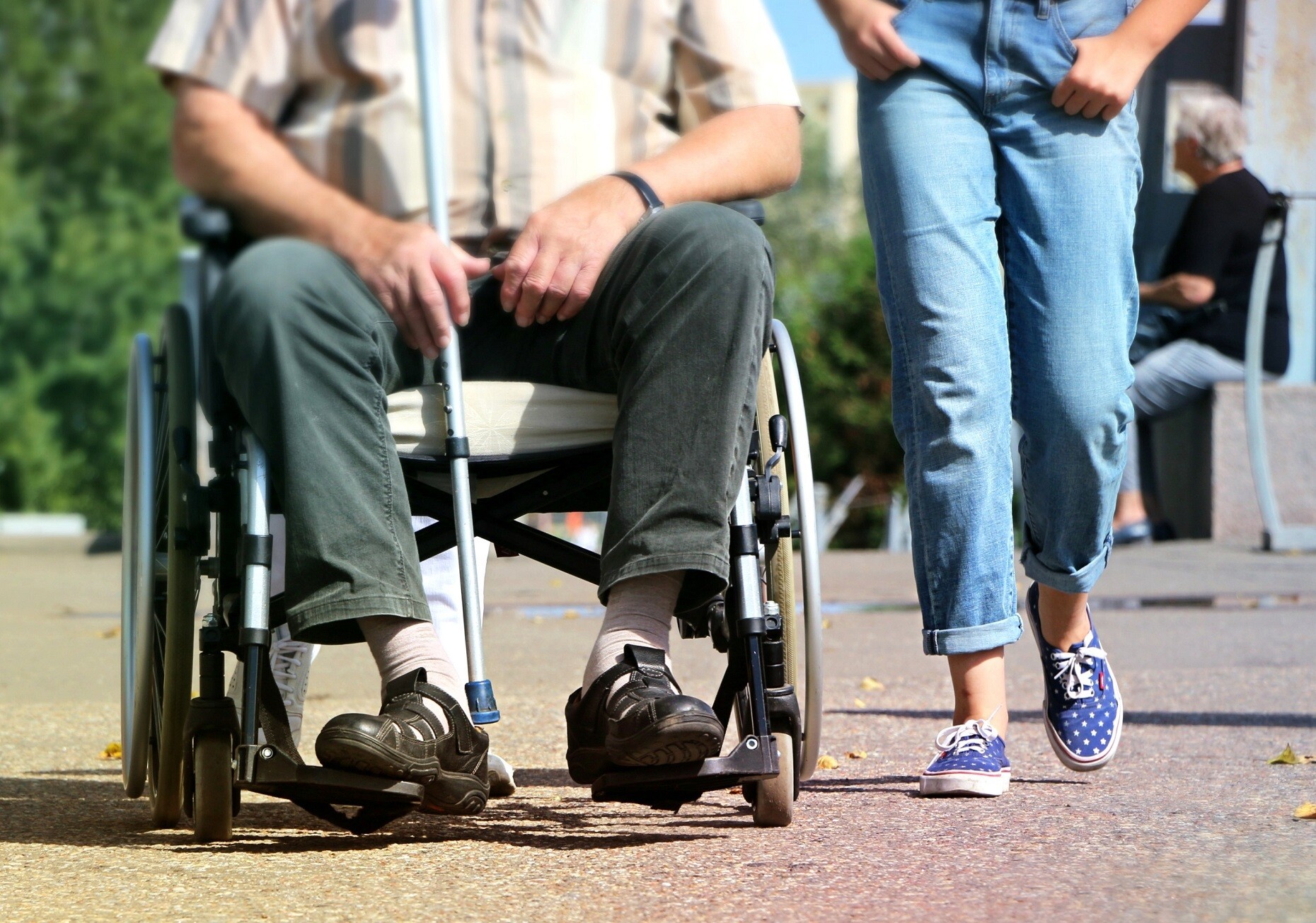 Ein pflegebedürftiger Mann in gestreiftem Hemd sitzt im Rollstuhl, rechts neben ihm läuft eine weibliche Begleitperson, die Hände in den Jeanstaschen. Die Köpfe sind nicht zu sehen.
