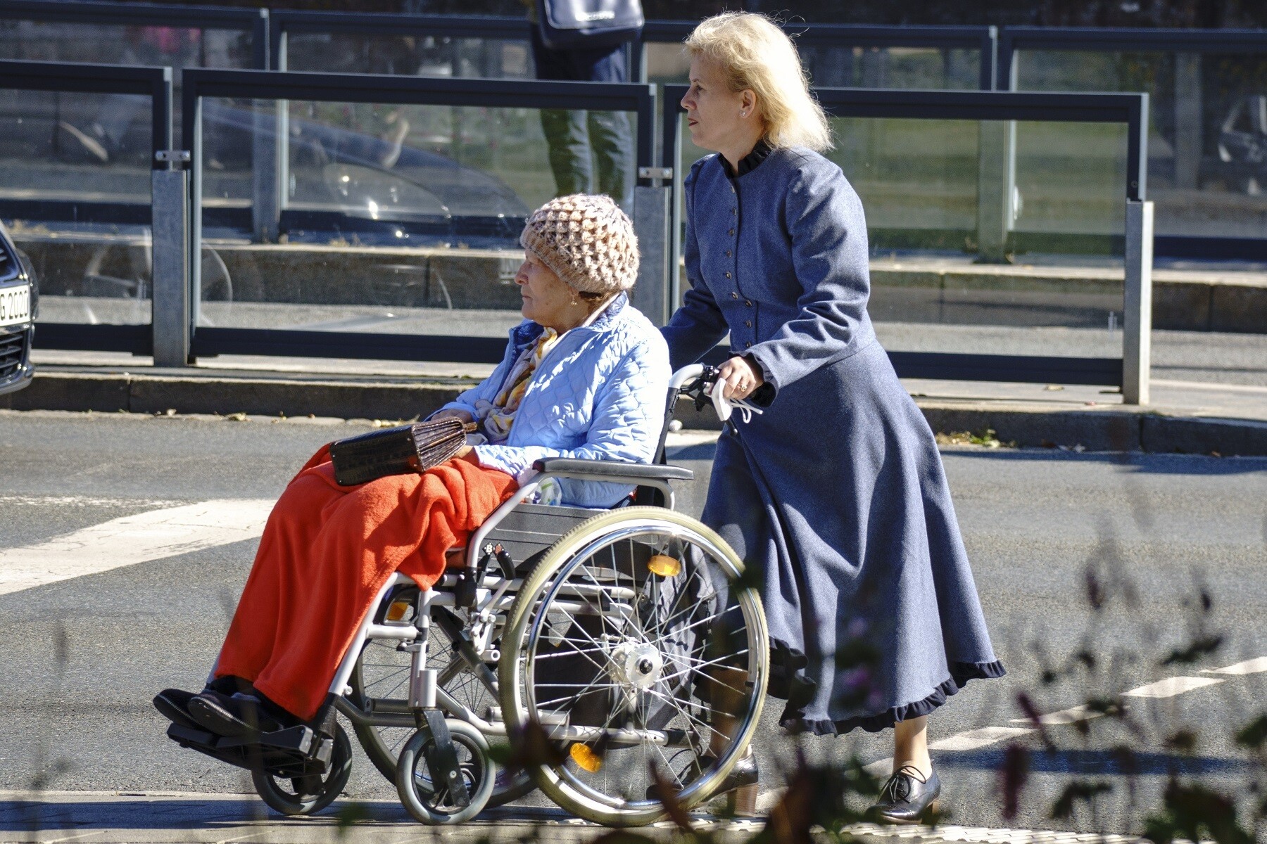 Une dame âgée est assise dans un fauteuil roulant. Elle porte un bonnet de laine beige et une veste bleu clair, ses jambes sont recouvertes d'une couverture rouge. Sa fille, une femme d'environ 50 ans aux cheveux blonds et au long manteau, la pousse sur le passage piéton.