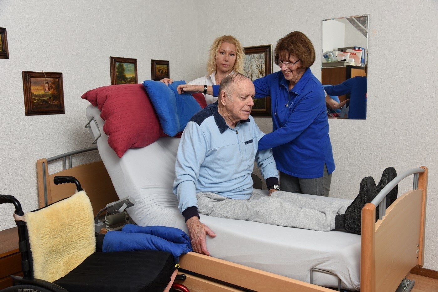 Zwei Frauen helfen einen älteren Mann zu Hause, sich im Bett aufzurichten. Neben dem Bett steht ein Rollstuhl bereit.
